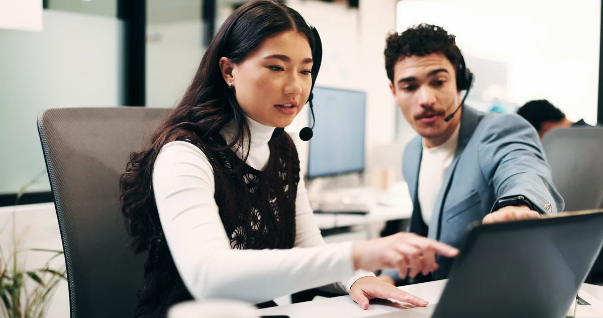 Two agency team members wearing headsets, working together at a laptop in an office