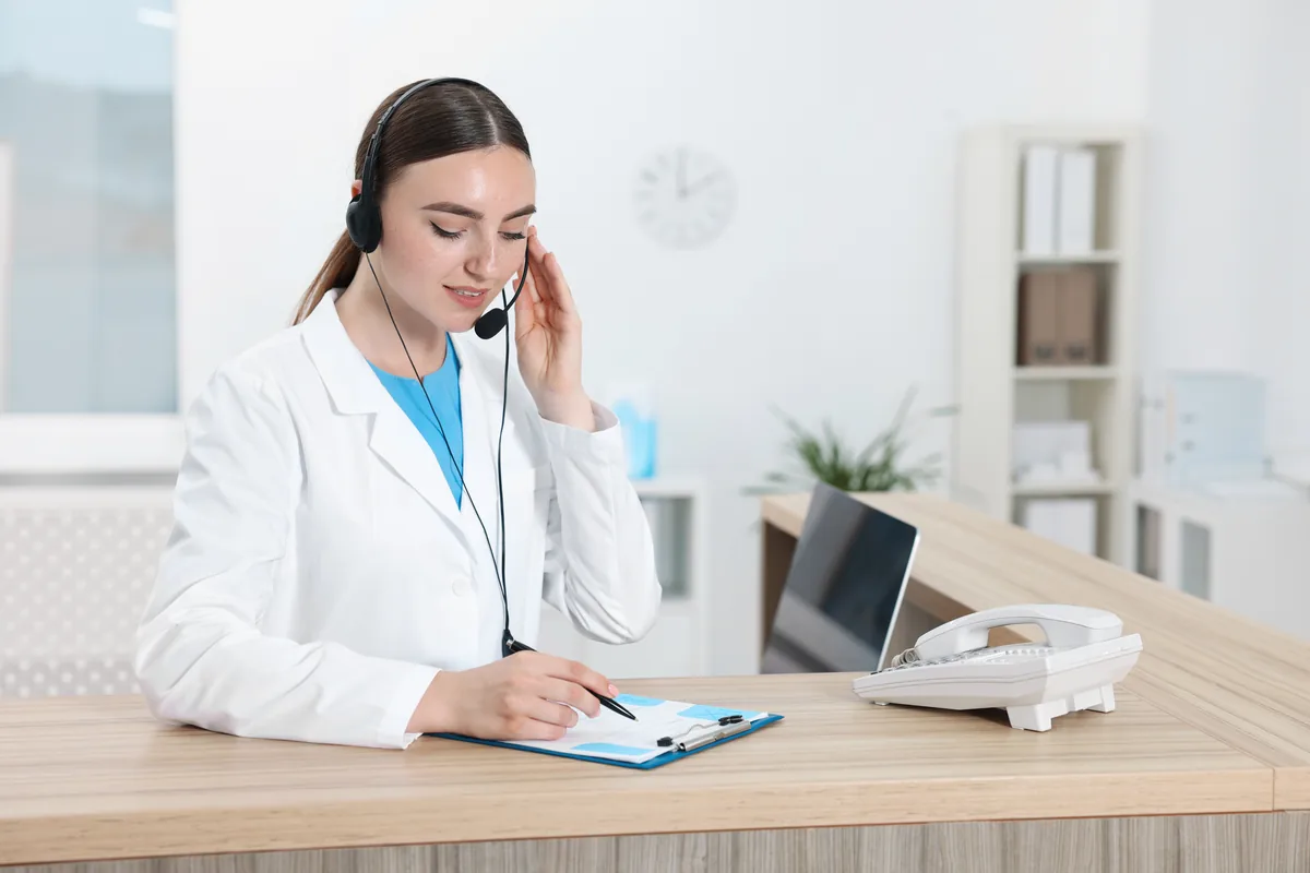 Receptionist in a white coat wearing a headset, taking a patient call at a front desk